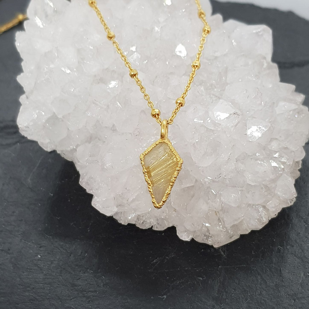 rutilated quartz rough diamond-shaped necklace set in 24ct gold with 45cm chain, handmade in the UK, displayed on a quartz stone